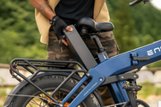 A person in camouflage clothing holds a large black rectangular removable battery mounted on a blue folding electric bicycle. 
