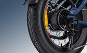 Close-up of a black bicycle wheel with a black tire, silver spokes, a black cassette, a silver chain, and a bright orange rear shock absorber against a blue gradient background.