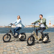 Two people riding electric bikes by a waterfront with a cityscape in the background